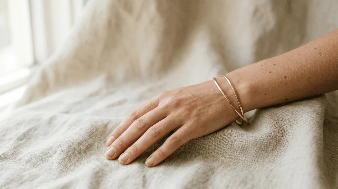 Close-up of model wearing two thin gold bangles on wrist resting against linen
