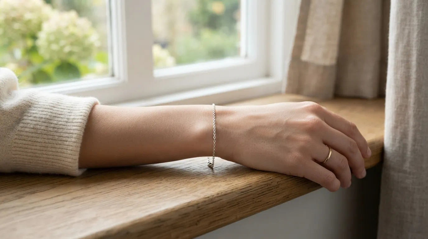 UGC close-up lifestyle photo of a model's wrist wearing a thin silver chain bracelet next to a wooden windowsill